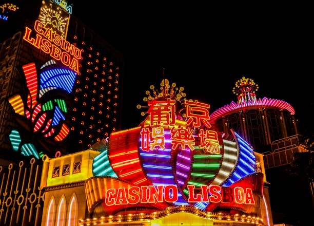 Bright neon lights and colorful signage adorn the exterior of Casino Lisboa, creating a vibrant and lively atmosphere. The façade features bold, multicolored designs with a mix of English and Chinese characters prominently displayed. The lights form intricate patterns and shapes, contributing to the energetic ambiance of the scene.
